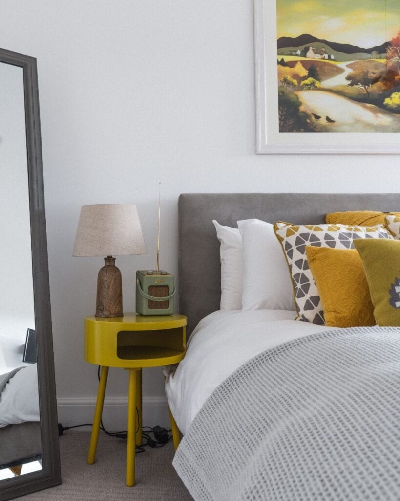 Grey upholstered bed with mustard cushions beside a yellow bedside table and landscape painting