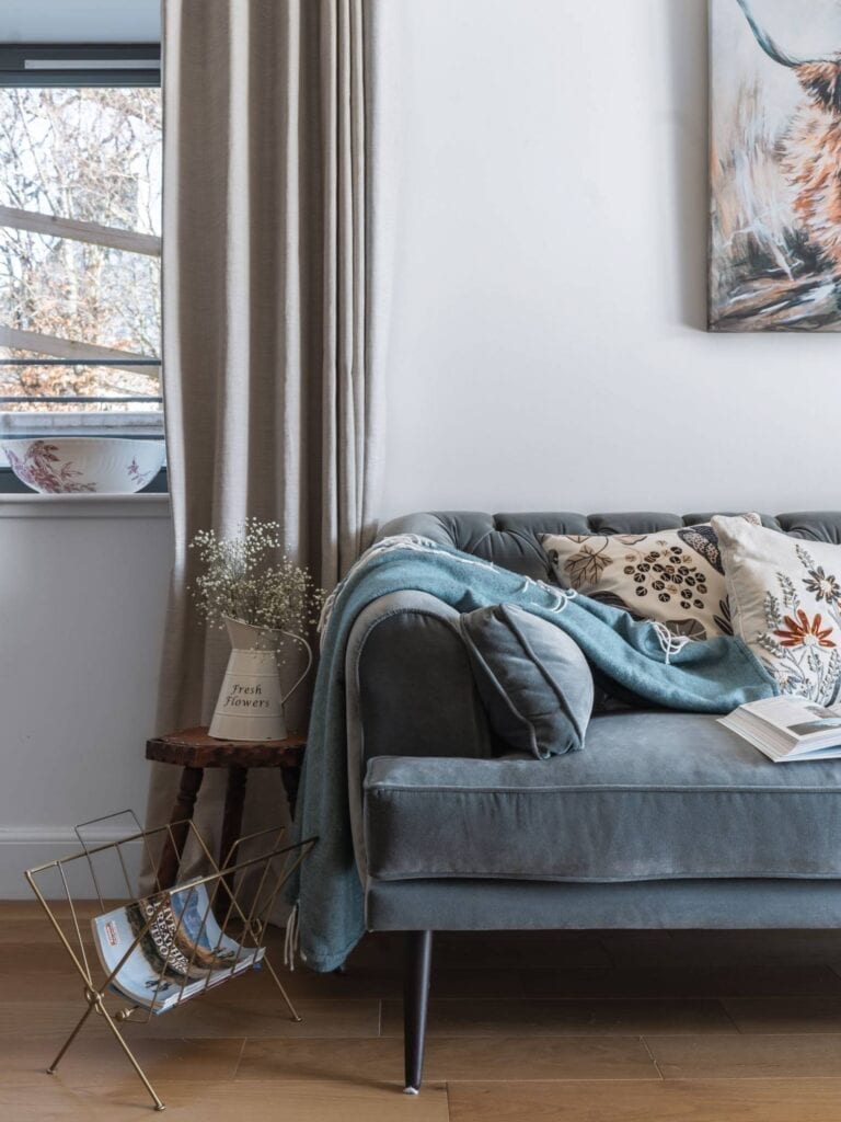 Grey velvet sofa with cushions and throw beside a "Fresh Flowers" jug