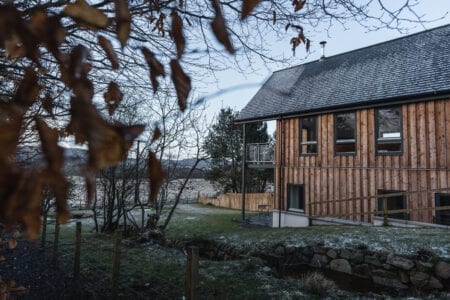 Timber-clad lodge with balcony overlooking a loch, framed by bare branches