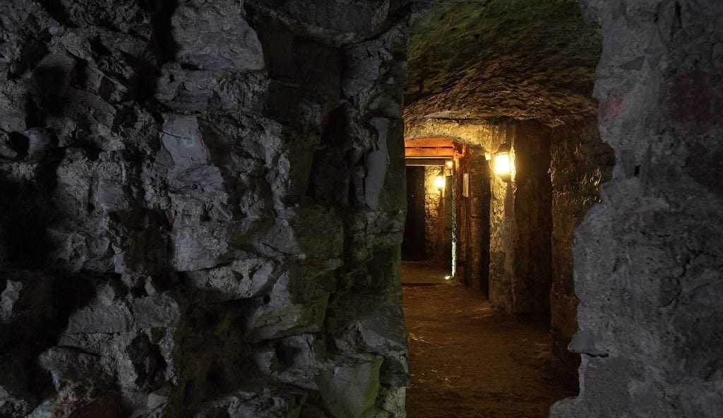 Stone-vaulted underground passage lit by wall lamps, rough rock walls either side