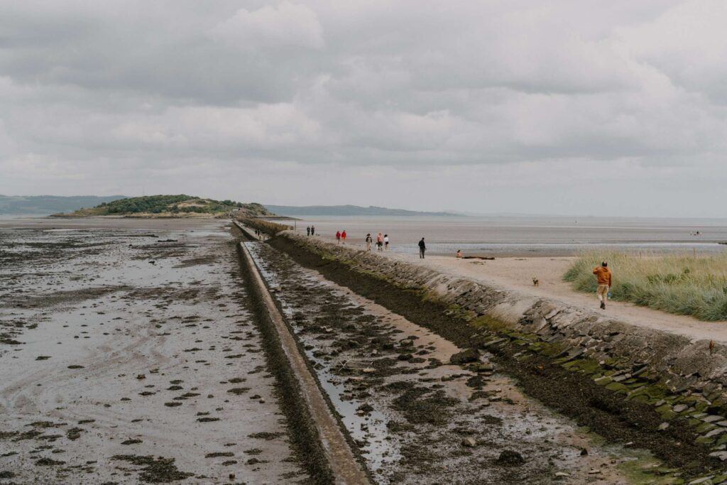 Walkers follow a causeway path between mudflats and sandy beach under grey skies