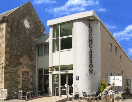 Modern entrance with vertical "Lochcarron" lettering beside a stone church building