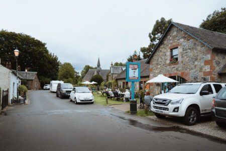 Village street with parked cars, a stone building and church spire beyond
