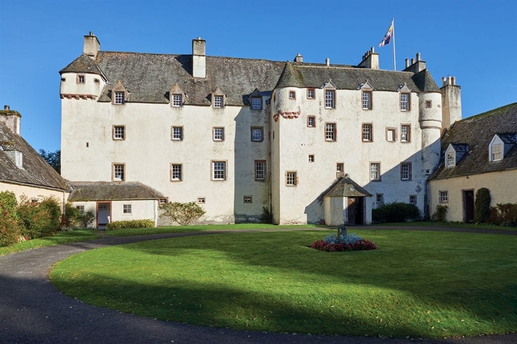 White harled tower house with turrets overlooking a lawn and flowerbed