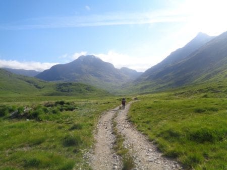 Walker on a gravel track through a glen towards distant peaks
