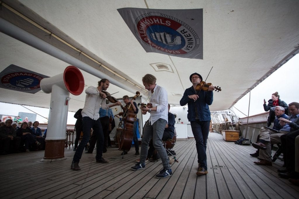 Musicians play fiddles and double bass on a ship's wooden deck