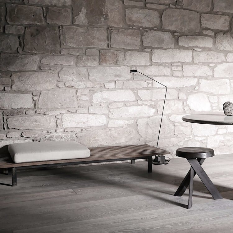 A wooden daybed with a bolster cushion sits before a stone wall, an angled floor lamp and a round table with a stool nearby.