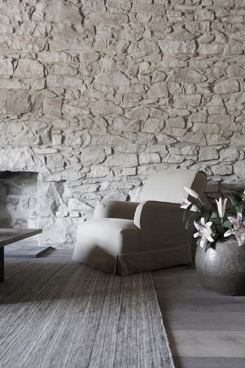 A cream upholstered armchair stands before a rough stone fireplace, a vase of lilies and a rug completing the room.