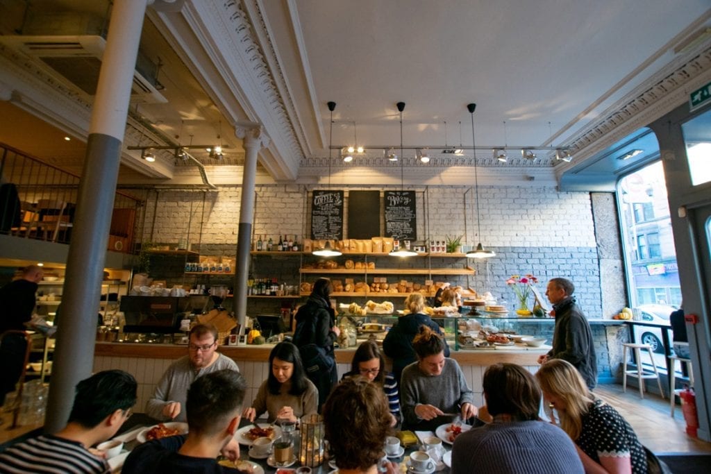 The busy café under high ceilings, counter and shelves behind the tables.