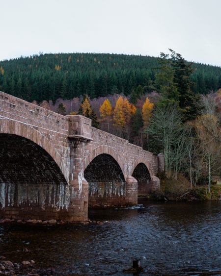 Stone arched bridge crossing a river below forested hills in autumn