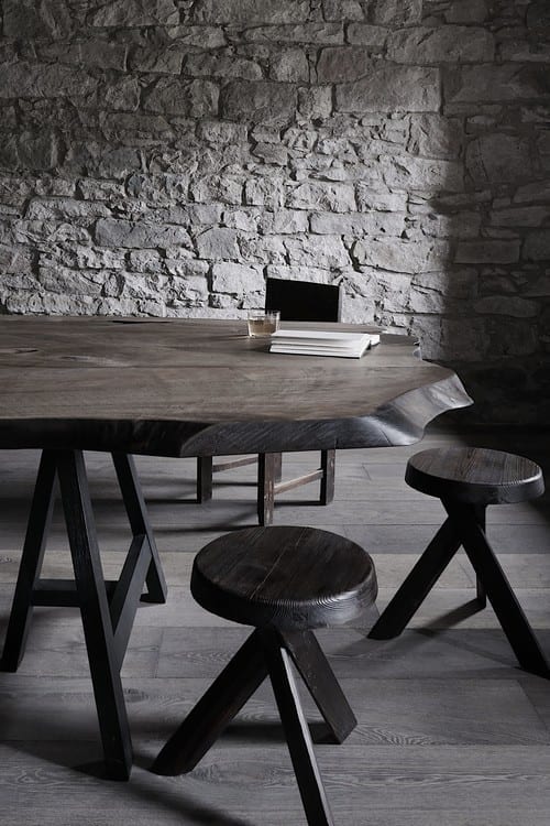 A live-edge wooden table with a book and glass stands before a stone wall, two turned wooden stools nearby.