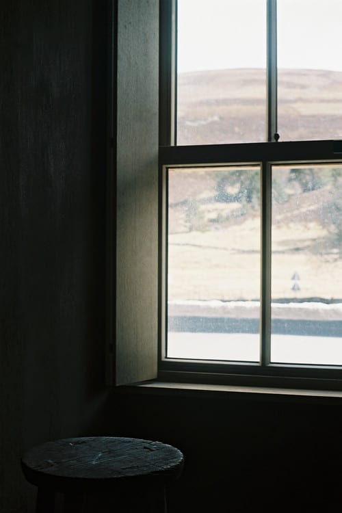 A wooden stool sits before a sash window looking out over moorland, the room around it in shadow.