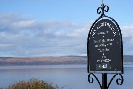Wrought-iron sign for The Lighthouse Restaurant beside a calm loch