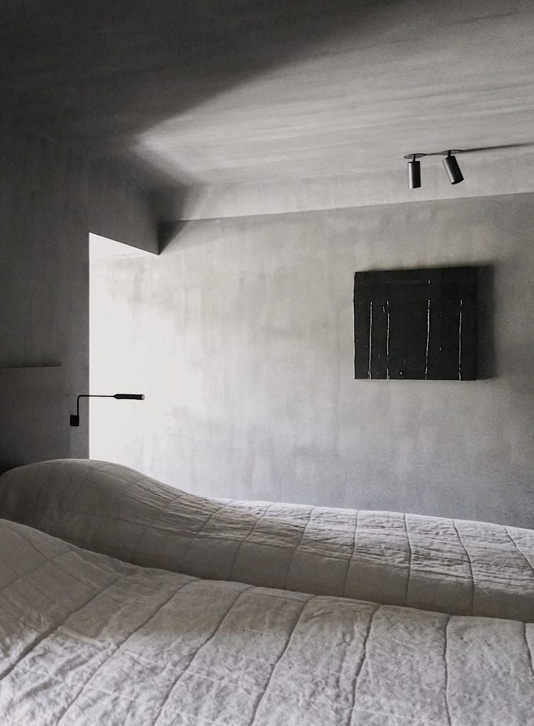 A quilted bed sits in a dimly lit room beneath a small dark painting, a reading lamp mounted on the wall to the left.