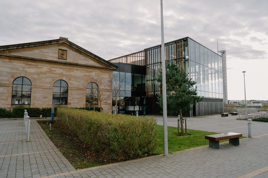 Sandstone building beside a modern glass extension with a wooden bench nearby