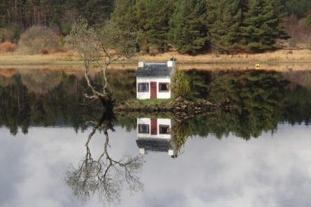 White cottage with red door on a small island, mirrored in still water