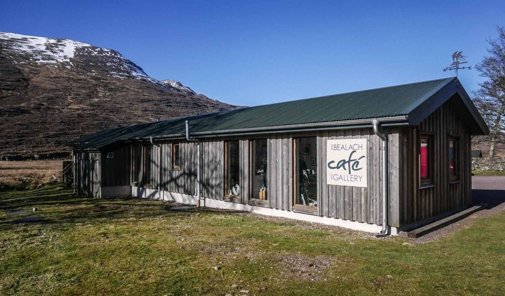 Timber-clad Bealach Café and Gallery below snow-streaked mountains under blue sky