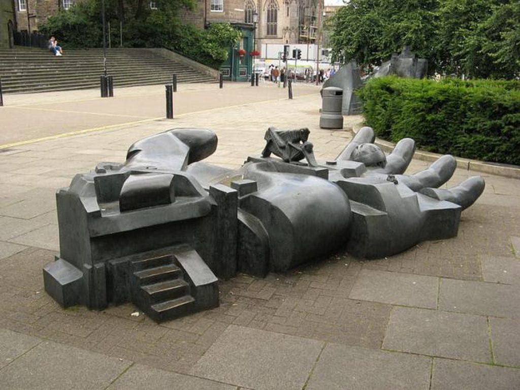 Abstract bronze sculpture on a paved plaza, with stone steps and trees behind