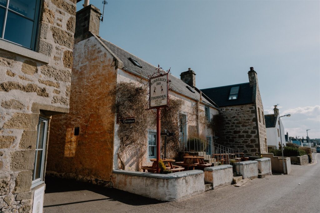 Stone cottages line a quiet street, with a hanging pub sign and outdoor tables