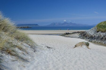 Sandy dunes frame a beach with distant hills across calm blue water