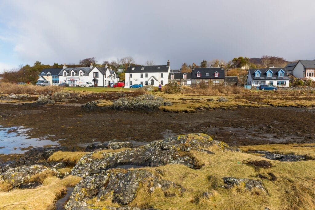 White houses along the shoreline at low tide, seaweed-covered rocks in foreground