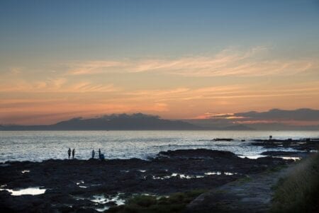 Figures on rocky shoreline silhouetted against pink sunset over distant hills