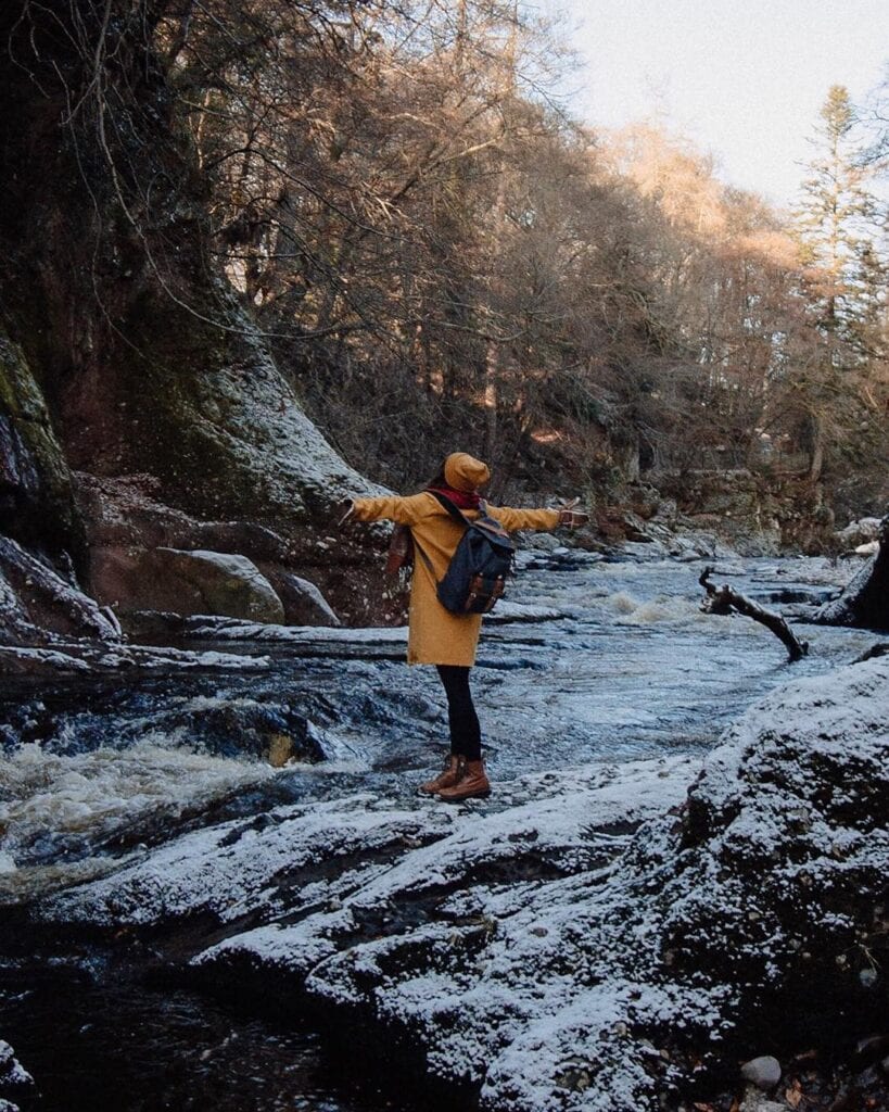 Walker in yellow coat balances on frosty rocks beside a river