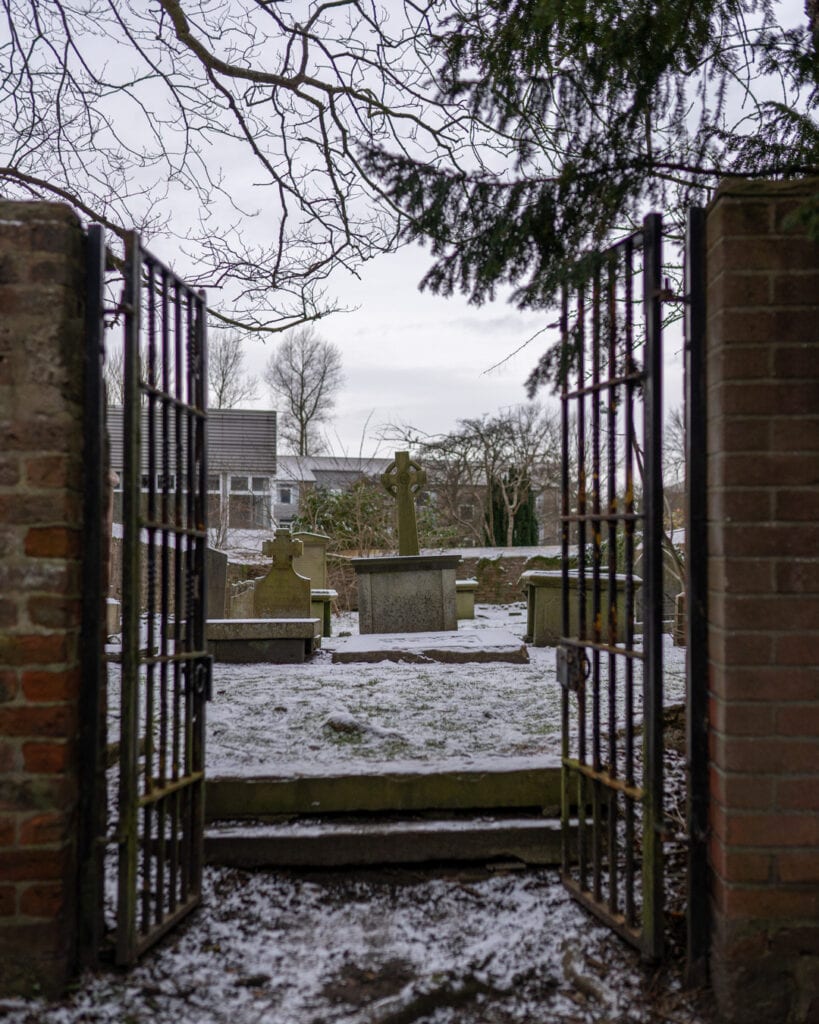 Open iron gates lead into a snow-dusted graveyard with a Celtic cross headstone