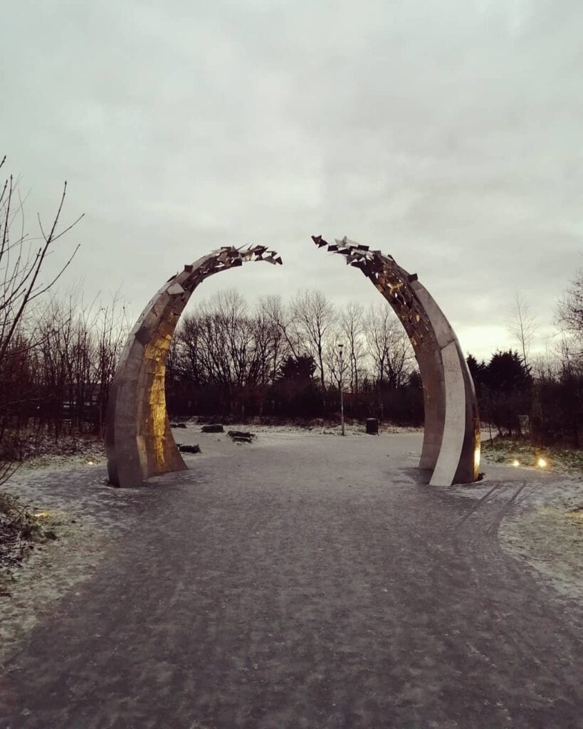 Two curved steel sculptures forming a gateway over a path at Cuningar Loop, frost on the ground under a grey sky.
