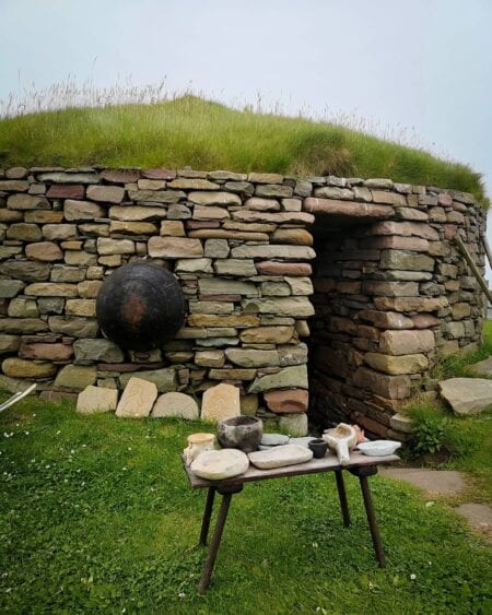 A reconstructed Iron Age house at Old Scatness, its turf roof and doorway, a cooking pot outside.