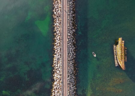 A barrier of stone blocks and its road from directly above, a rusting blockship in the green water beside it.