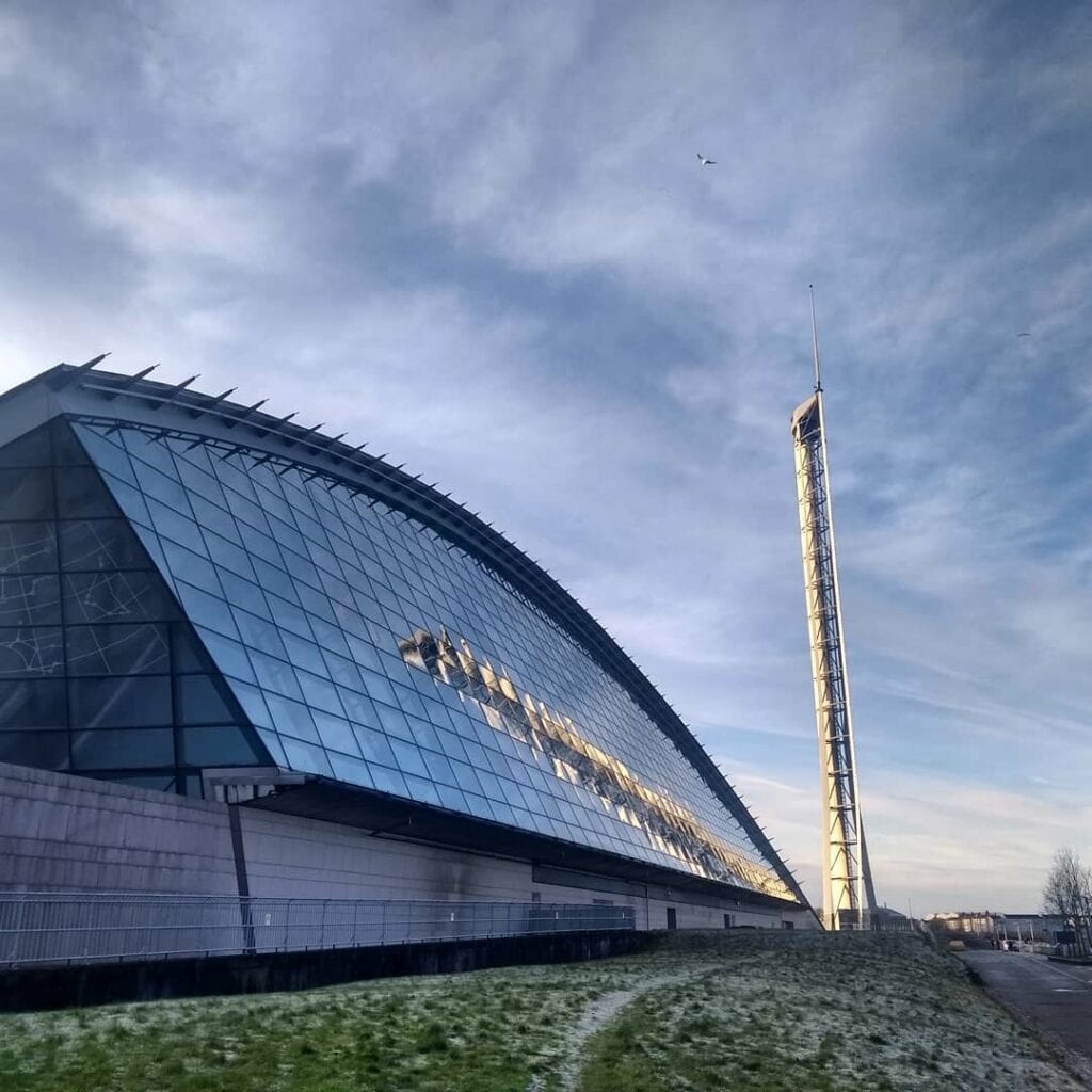 The curved glass end of the Glasgow Science Centre and the Glasgow Tower against a pale sky.