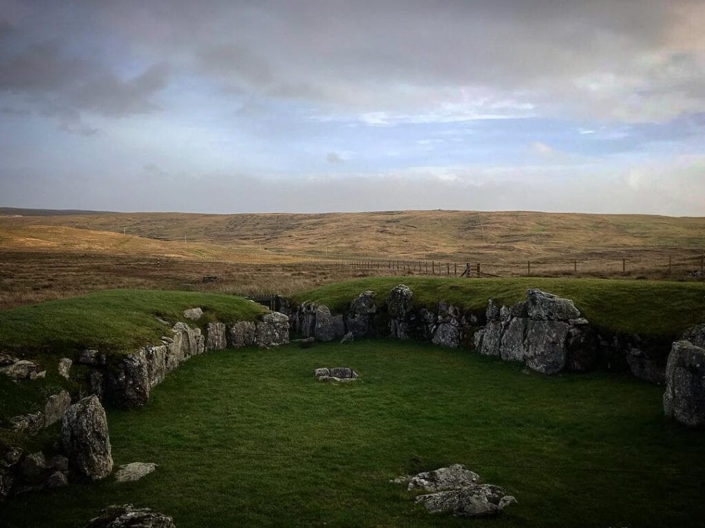 The heel-shaped stone walls of Stanydale Temple in the moorland under a cloudy sky.
