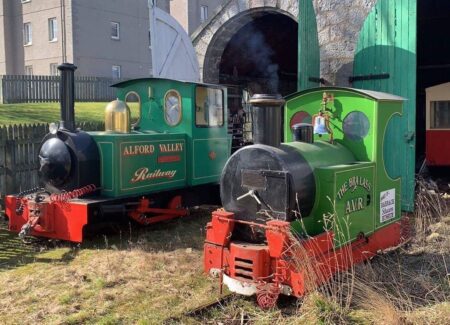 Two little green engines of the Alford Valley Railway with red buffers, outside the engine shed.