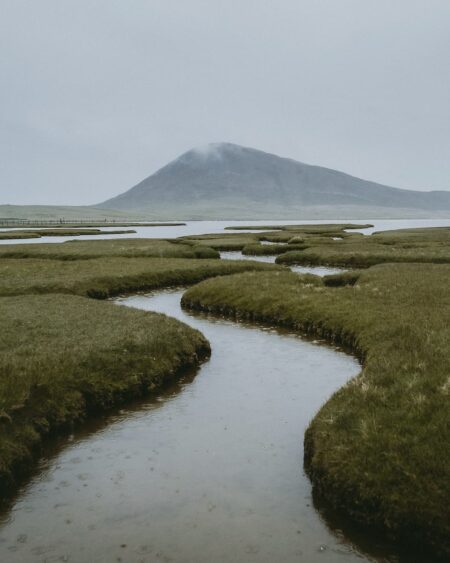 A winding tidal channel cuts through saltmarsh grass beneath a cloud-capped hill