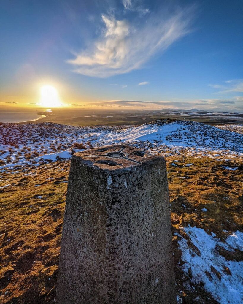 Trig point on a snow-patched hilltop overlooking the coast at sunset