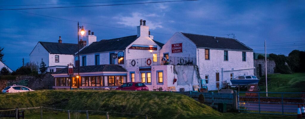 White-painted inn with lit windows overlooking a grassy slope at dusk