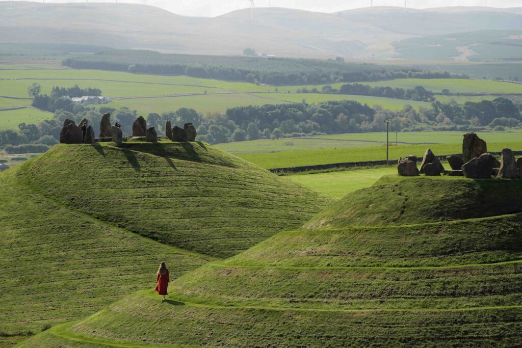 Woman in red walking grassy hills below a hilltop stone circle