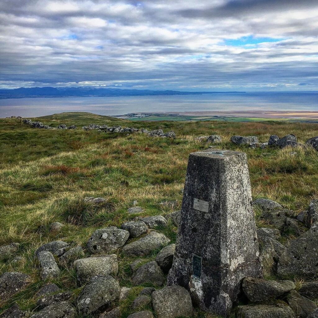 Trig point on a grassy summit overlooking a distant coastline and sea