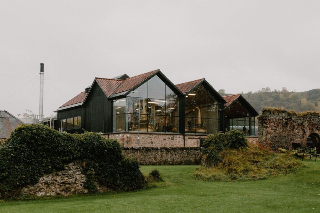 Glass-fronted distillery buildings beside a ruined stone wall and chimney