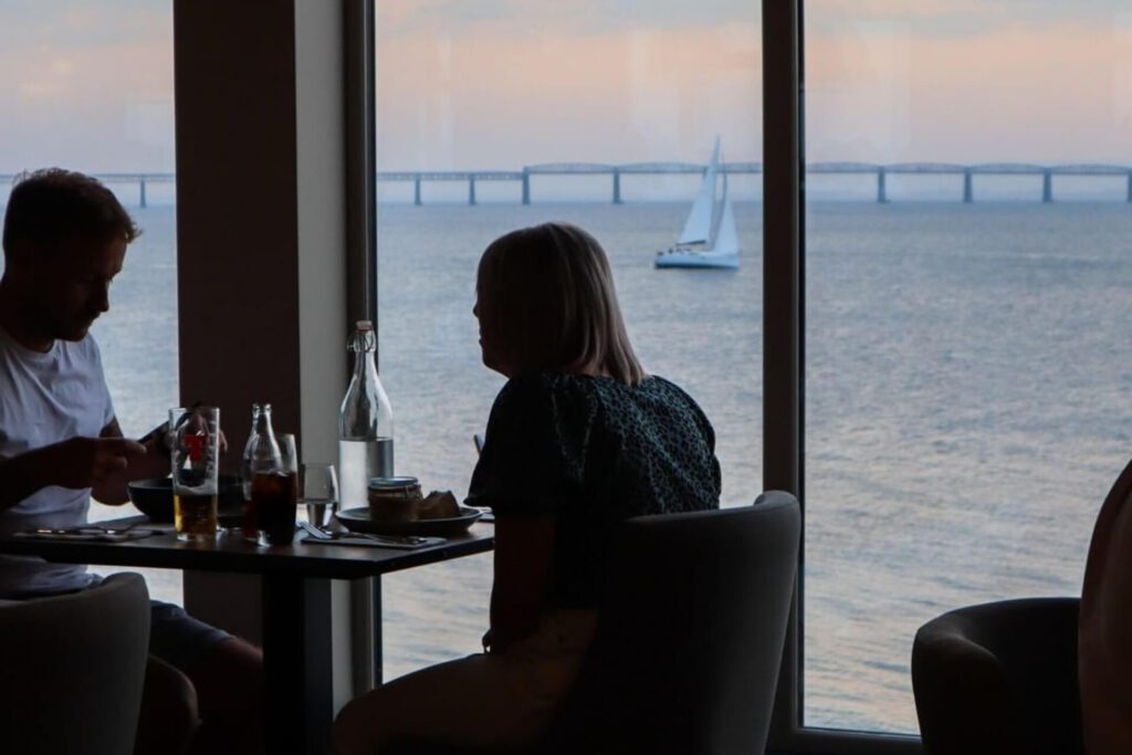 Diners at a window table overlook a sailing boat and distant bridge