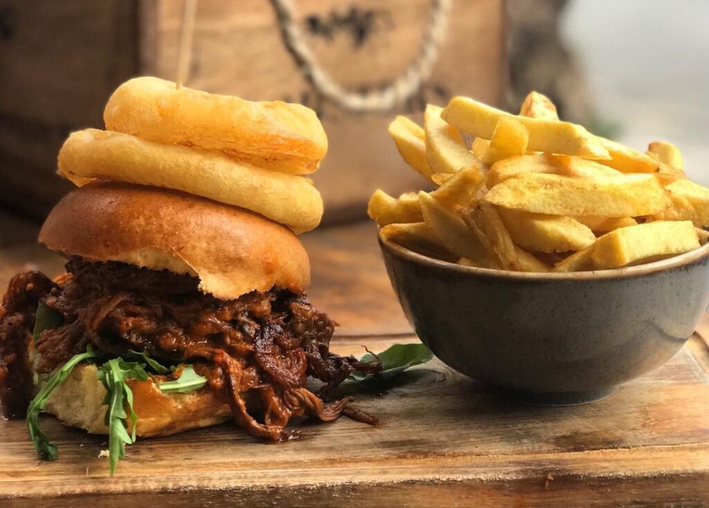 A pulled beef burger topped with onion rings, and a bowl of chips.