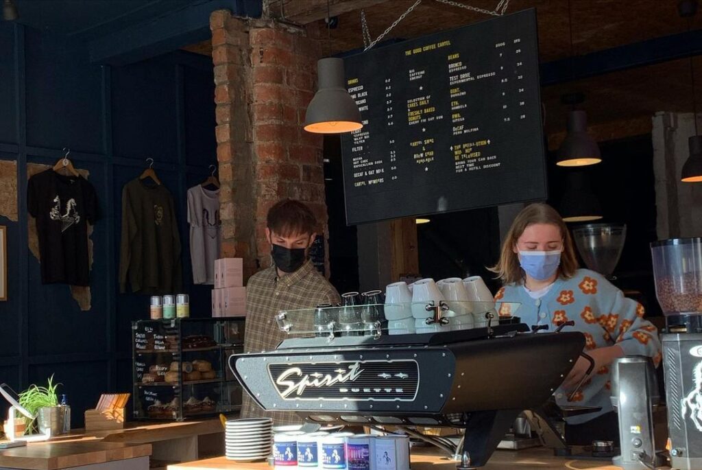 Two baristas behind the Spirit espresso machine under the drinks board.