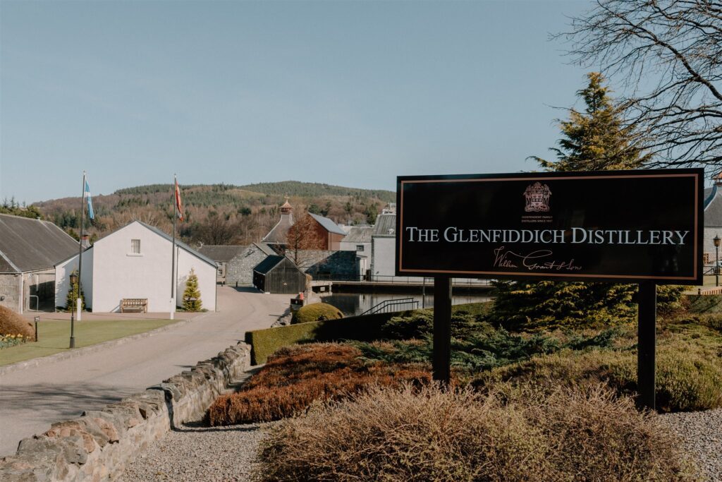 The Glenfiddich Distillery sign beside whitewashed buildings and a stone wall