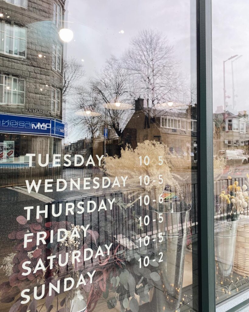 Shop window listing opening hours, Tuesday to Sunday, reflecting the street outside
