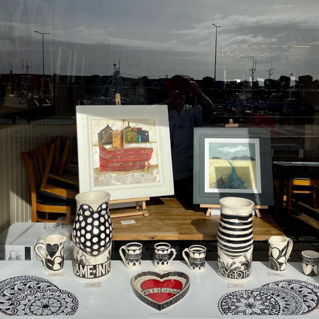 Shop window display of framed paintings, patterned ceramics and heart-shaped dish, harbour reflected in glass