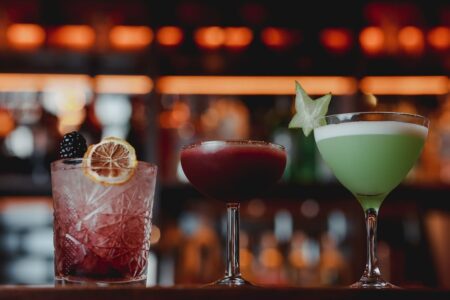 Three cocktails on a bar, garnished with blackberry, lemon and starfruit