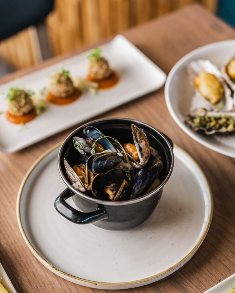 Mussels in a black pot beside breaded croquettes and dressed oysters on a wooden table