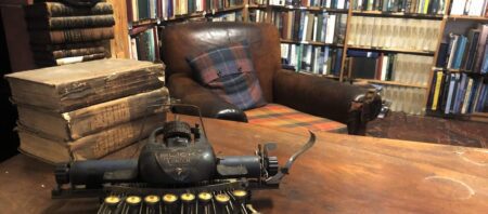 Vintage typewriter and stacked leather books beside a tartan-cushioned armchair in a library