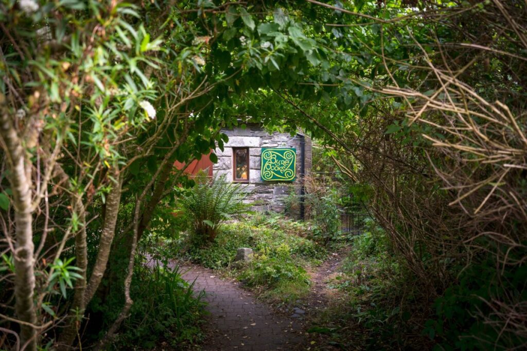 Stone cottage with a green Celtic knot sign, seen through a leafy tunnel